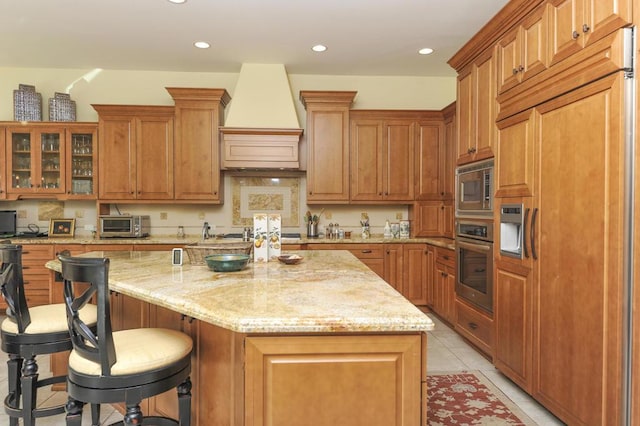 kitchen featuring premium range hood, light tile patterned flooring, an island with sink, built in appliances, and light stone countertops