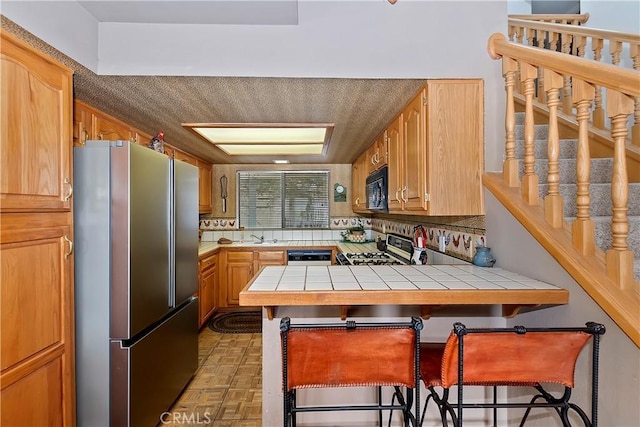 kitchen featuring tile countertops, dishwasher, stainless steel fridge, a kitchen bar, and range