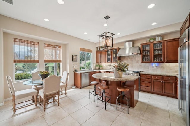 kitchen featuring decorative light fixtures, a kitchen island, stainless steel appliances, light stone countertops, and wall chimney range hood