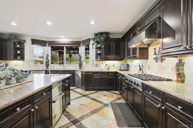 kitchen featuring tasteful backsplash, light stone counters, dark brown cabinetry, stainless steel appliances, and wall chimney range hood