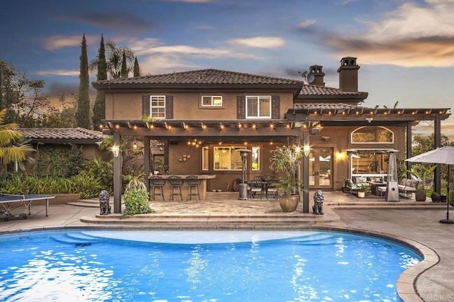 pool at dusk featuring exterior bar, a pergola, and a patio area