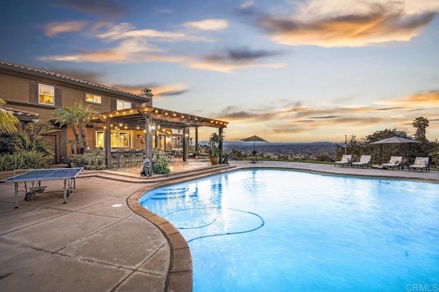 pool at dusk featuring a pergola, an outdoor bar, and a patio area