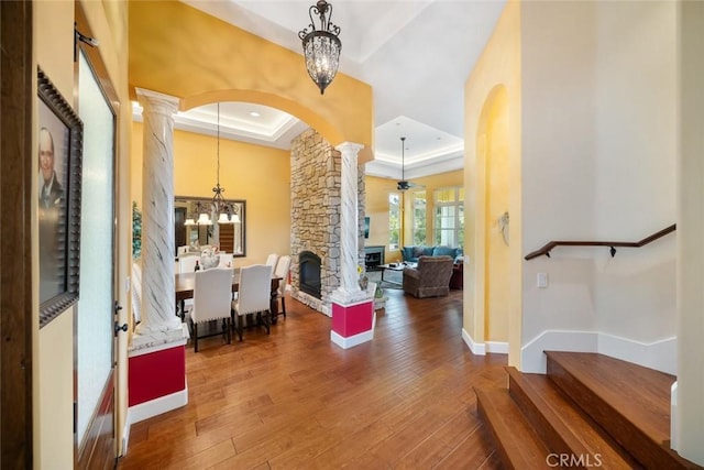 foyer entrance with hardwood / wood-style flooring, decorative columns, a stone fireplace, a raised ceiling, and a chandelier