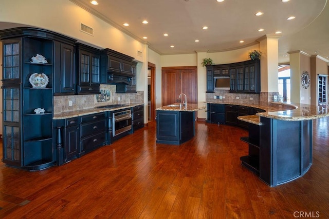 kitchen featuring appliances with stainless steel finishes, dark hardwood / wood-style floors, a kitchen island with sink, kitchen peninsula, and light stone countertops