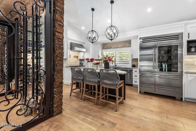 kitchen with wall chimney range hood, a kitchen breakfast bar, built in appliances, white cabinets, and decorative backsplash