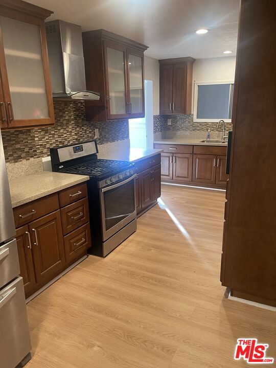 kitchen with wall chimney range hood, sink, stainless steel range with gas stovetop, tasteful backsplash, and light hardwood / wood-style floors