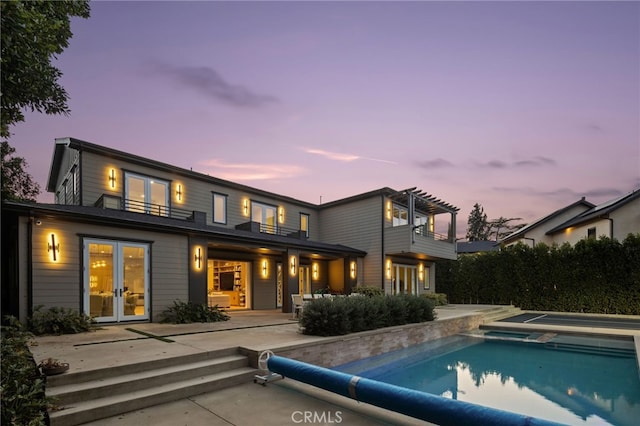 back house at dusk featuring french doors, a patio area, a balcony, a covered pool, and a pergola