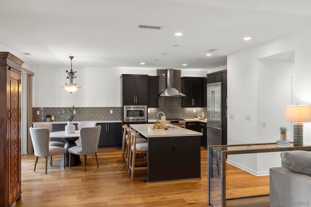 kitchen with hanging light fixtures, a kitchen island with sink, built in appliances, wall chimney exhaust hood, and light hardwood / wood-style flooring