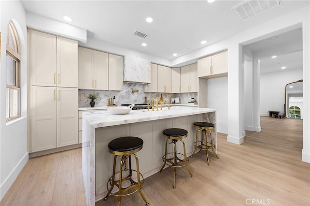 kitchen featuring an island with sink, a kitchen breakfast bar, decorative backsplash, light stone countertops, and light wood-type flooring