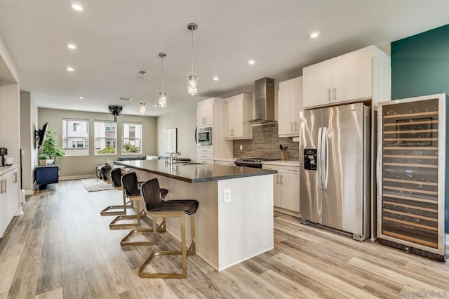 kitchen with decorative light fixtures, appliances with stainless steel finishes, an island with sink, beverage cooler, and wall chimney range hood