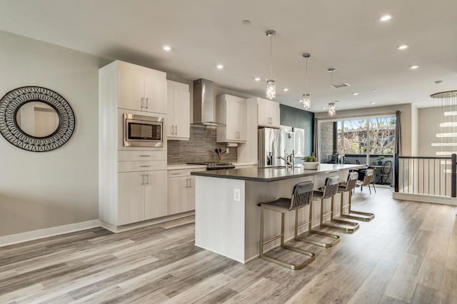 kitchen featuring wall chimney exhaust hood, decorative light fixtures, appliances with stainless steel finishes, a kitchen island with sink, and backsplash