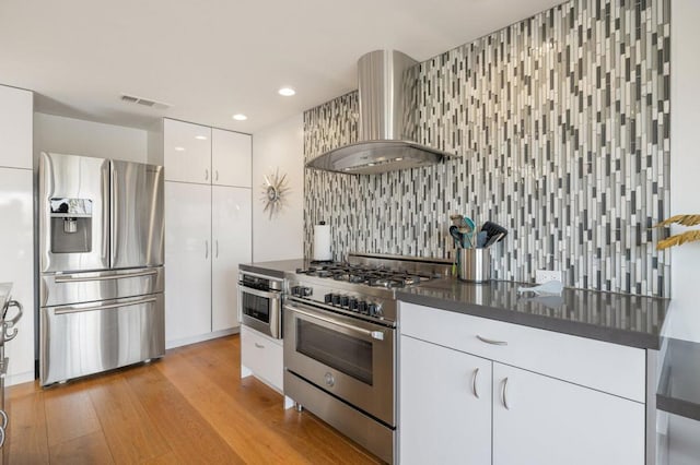 kitchen with wall chimney range hood, appliances with stainless steel finishes, white cabinetry, tasteful backsplash, and light hardwood / wood-style floors