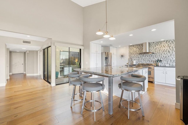 kitchen featuring light hardwood / wood-style flooring, hanging light fixtures, appliances with stainless steel finishes, white cabinets, and wall chimney range hood