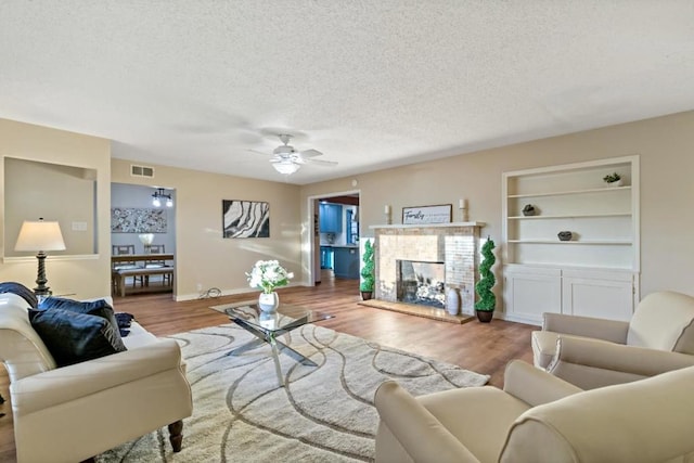 living room featuring a textured ceiling, light wood-type flooring, built in features, ceiling fan, and a fireplace