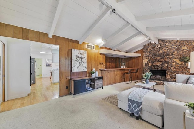 carpeted living room with a stone fireplace, vaulted ceiling with beams, and wooden walls