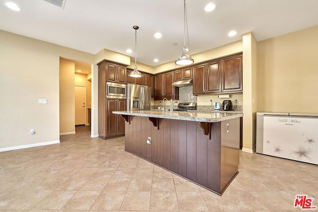 kitchen featuring appliances with stainless steel finishes, hanging light fixtures, a kitchen breakfast bar, light stone countertops, and a center island with sink