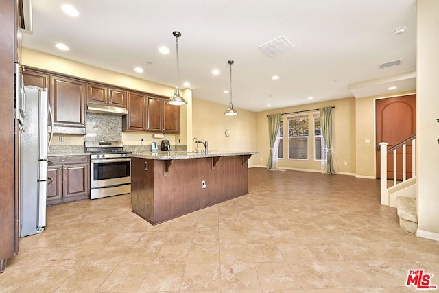 kitchen with a breakfast bar area, decorative light fixtures, a center island with sink, stainless steel appliances, and light stone countertops