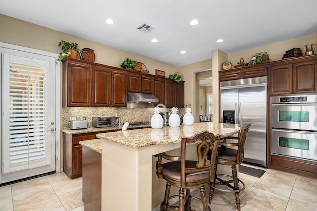 kitchen with a kitchen bar, light stone counters, appliances with stainless steel finishes, an island with sink, and backsplash