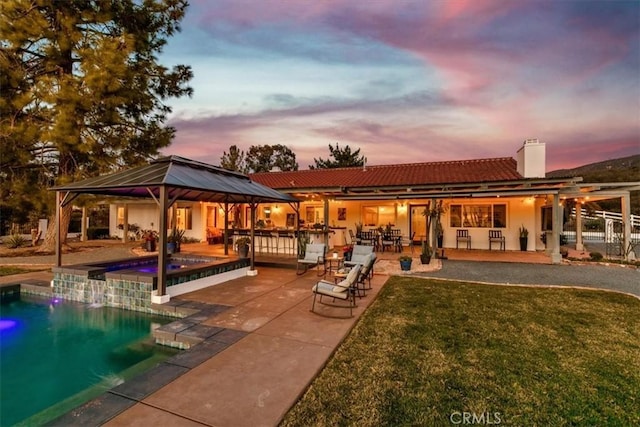 back house at dusk featuring a gazebo, a swimming pool with hot tub, a yard, and a patio