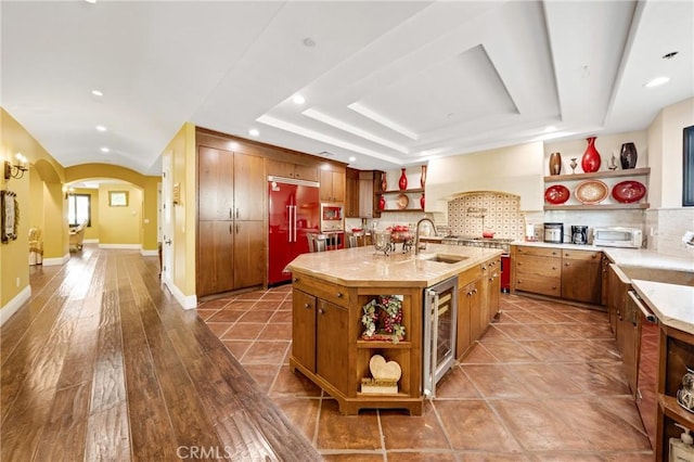 kitchen featuring built in refrigerator, wine cooler, a tray ceiling, a center island with sink, and decorative backsplash