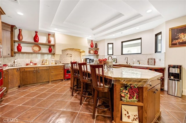 kitchen featuring premium range hood, dark tile patterned flooring, a kitchen bar, a kitchen island with sink, and a tray ceiling