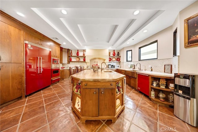 kitchen with sink, backsplash, built in appliances, an island with sink, and a raised ceiling