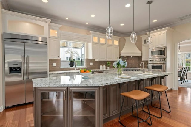 kitchen featuring white cabinetry, a spacious island, built in appliances, decorative light fixtures, and custom exhaust hood