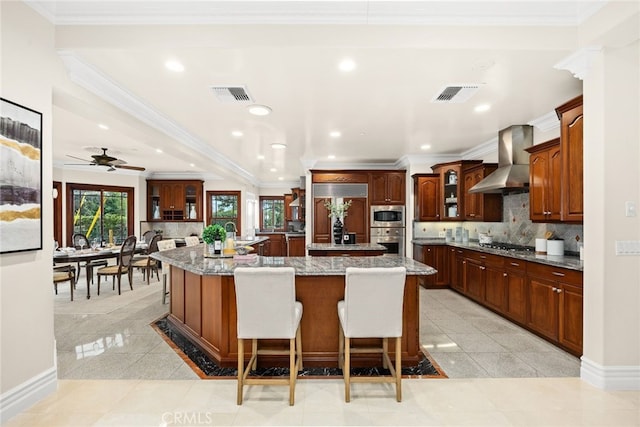 kitchen featuring a breakfast bar, an island with sink, decorative backsplash, stainless steel appliances, and wall chimney range hood