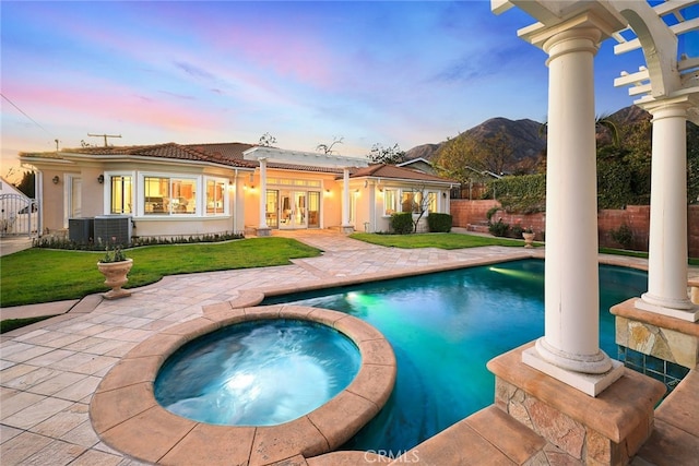 pool at dusk with french doors, an in ground hot tub, a yard, a mountain view, and a patio area