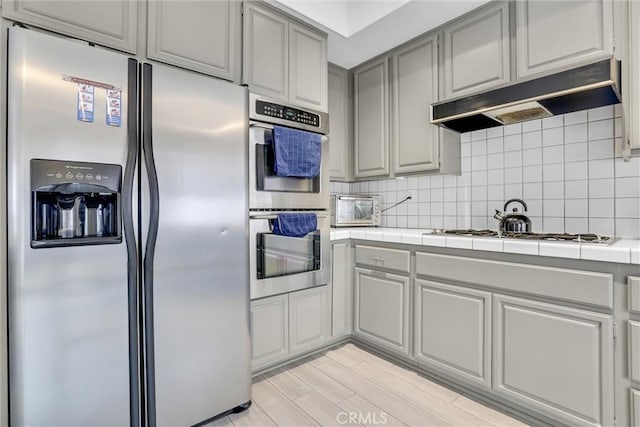 kitchen with tasteful backsplash, tile countertops, appliances with stainless steel finishes, gray cabinets, and light hardwood / wood-style floors