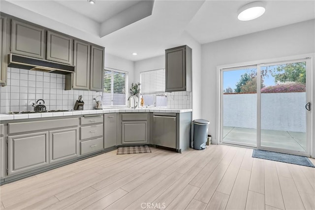 kitchen with gray cabinetry, stainless steel dishwasher, tile counters, and light wood-type flooring