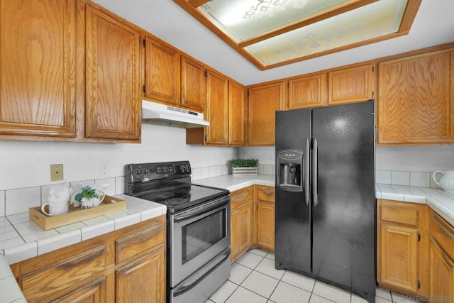 kitchen featuring light tile patterned floors, stainless steel range with electric stovetop, backsplash, tile counters, and black refrigerator with ice dispenser