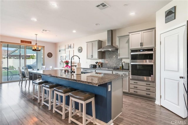 kitchen with a kitchen island with sink, gray cabinets, stainless steel appliances, and wall chimney exhaust hood