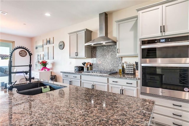 kitchen featuring wall chimney range hood, sink, stainless steel appliances, tasteful backsplash, and dark stone counters