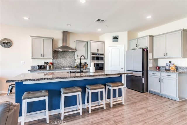 kitchen with refrigerator, a kitchen breakfast bar, stainless steel double oven, a kitchen island with sink, and wall chimney range hood