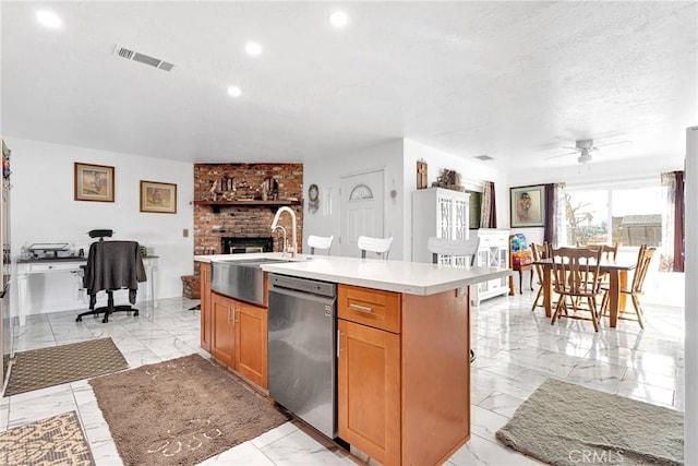 kitchen featuring sink, a brick fireplace, a center island with sink, stainless steel dishwasher, and ceiling fan