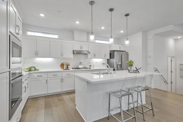 kitchen featuring appliances with stainless steel finishes, an island with sink, sink, white cabinets, and hanging light fixtures