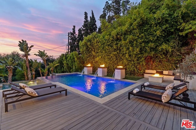 pool at dusk featuring a wooden deck, pool water feature, and an outdoor fire pit