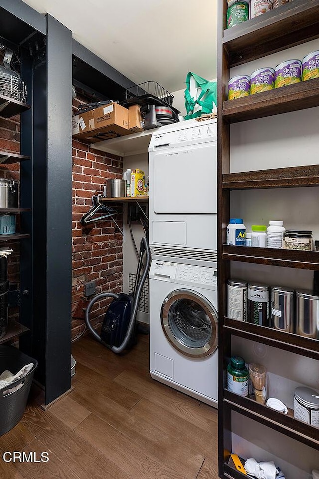 laundry area featuring brick wall, stacked washer / dryer, and wood-type flooring