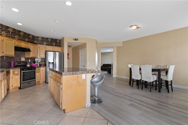 kitchen featuring dark stone countertops, stainless steel appliances, a center island, decorative backsplash, and light brown cabinets