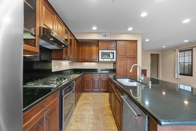 kitchen with sink, light tile patterned floors, appliances with stainless steel finishes, a kitchen island with sink, and dark stone counters