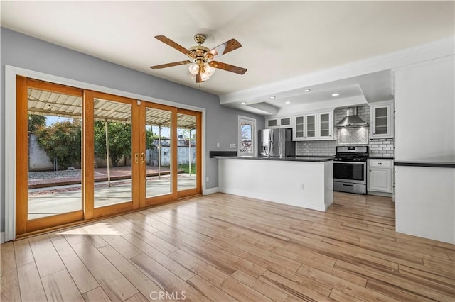 kitchen featuring white cabinets, decorative backsplash, kitchen peninsula, stainless steel appliances, and wall chimney range hood