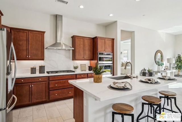 kitchen with wall chimney exhaust hood, a breakfast bar, sink, a center island with sink, and appliances with stainless steel finishes