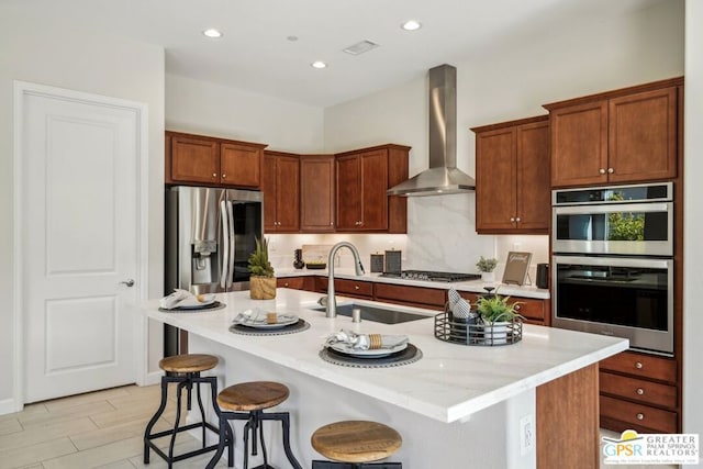 kitchen featuring appliances with stainless steel finishes, a kitchen island with sink, wall chimney range hood, and a kitchen bar