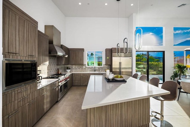 kitchen featuring a breakfast bar area, built in appliances, a kitchen island, decorative light fixtures, and wall chimney exhaust hood