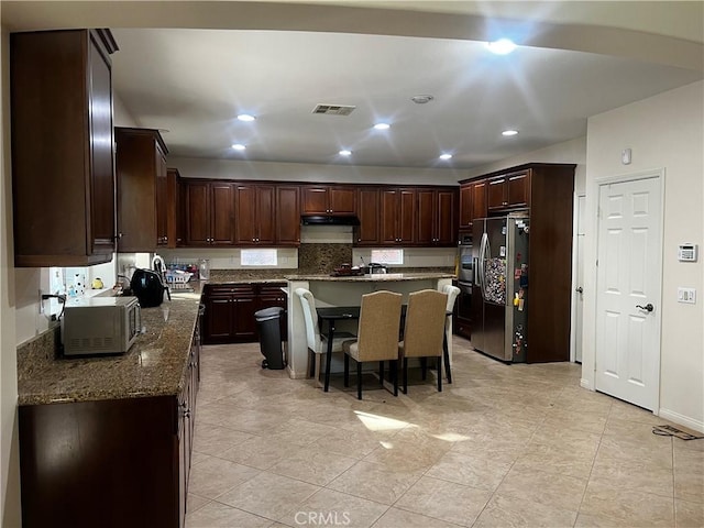 kitchen with dark stone countertops, stainless steel fridge, a kitchen bar, a center island, and dark brown cabinetry