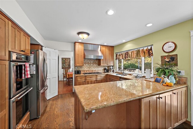 kitchen with wall chimney exhaust hood, tasteful backsplash, appliances with stainless steel finishes, kitchen peninsula, and light stone countertops