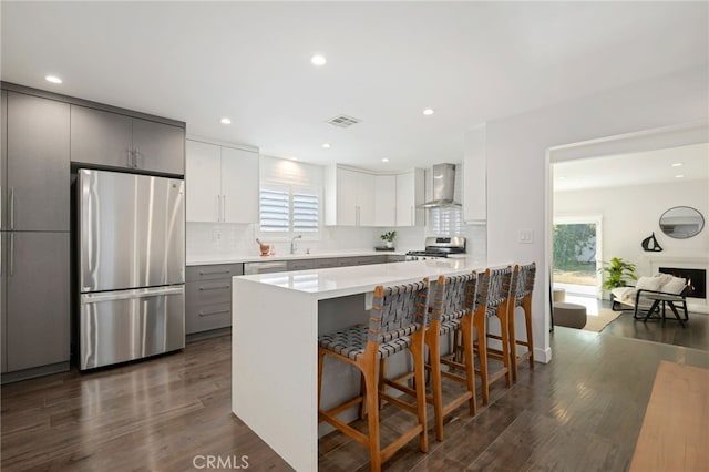 kitchen with gray cabinets, appliances with stainless steel finishes, a breakfast bar, a wealth of natural light, and wall chimney exhaust hood