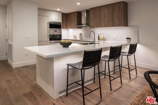 kitchen featuring wall chimney exhaust hood, sink, a breakfast bar area, kitchen peninsula, and double oven