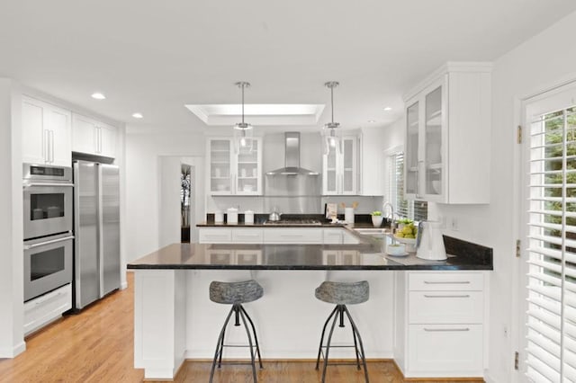 kitchen featuring a breakfast bar, white cabinetry, appliances with stainless steel finishes, pendant lighting, and wall chimney range hood